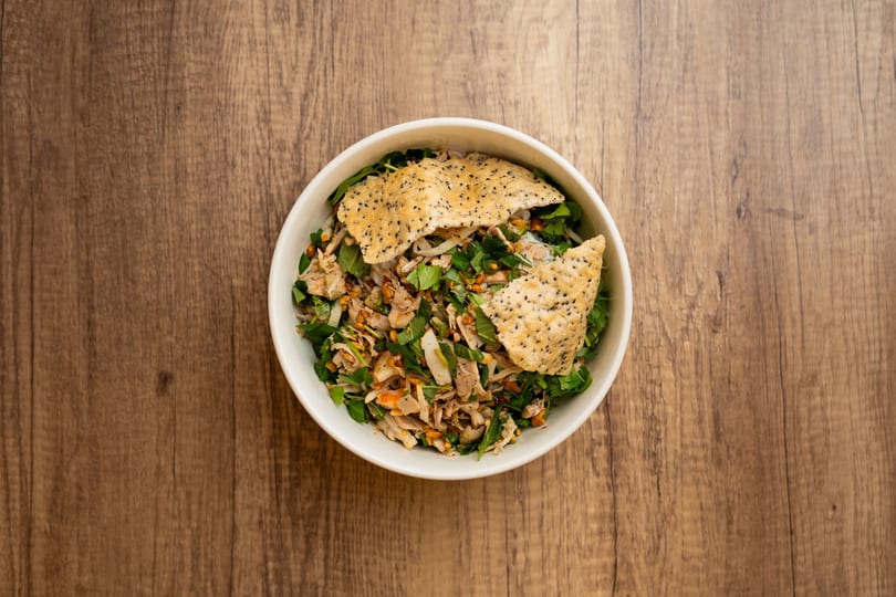 Overhead view of a Vietnamese chicken salad with herbs and sesame rice crackers in a white bowl.