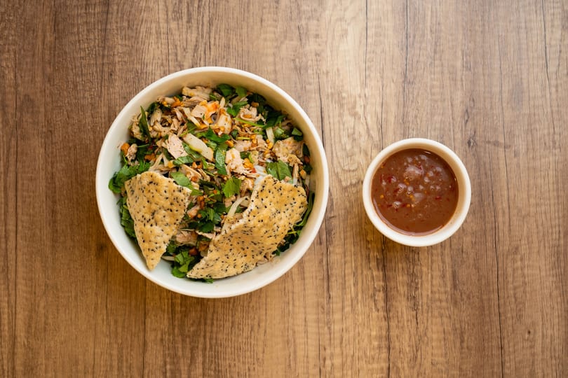 A bowl of Vietnamese chicken salad with herbs and crispy sesame crackers, next to a dipping sauce.