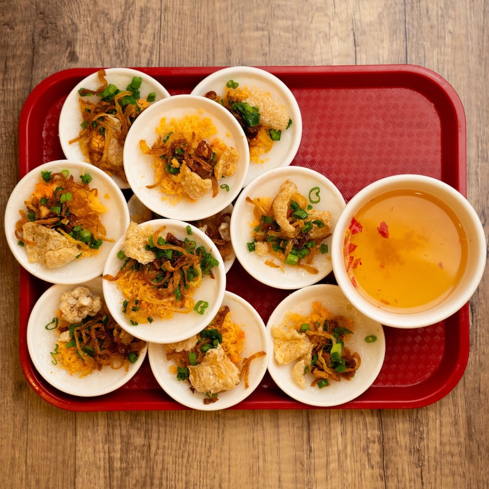 A red tray holds small white dishes of Vietnamese Banh Beo with crispy toppings and a dipping sauce.